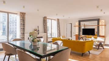 A modern living room with a glass dining table, yellow sofa, and a large TV.