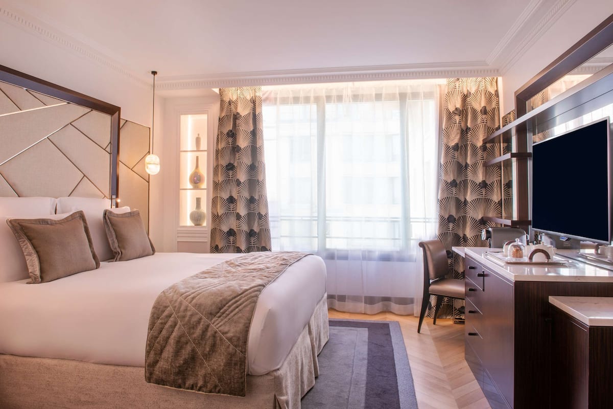 Habitación de hotel con una cama grande, un escritorio con televisión de pantalla plana y una ventana con cortinas translúcidas.