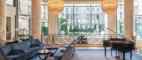 A spacious lobby with a grand piano, elegant seating, large chandeliers, and a view of the city through floor-to-ceiling windows.