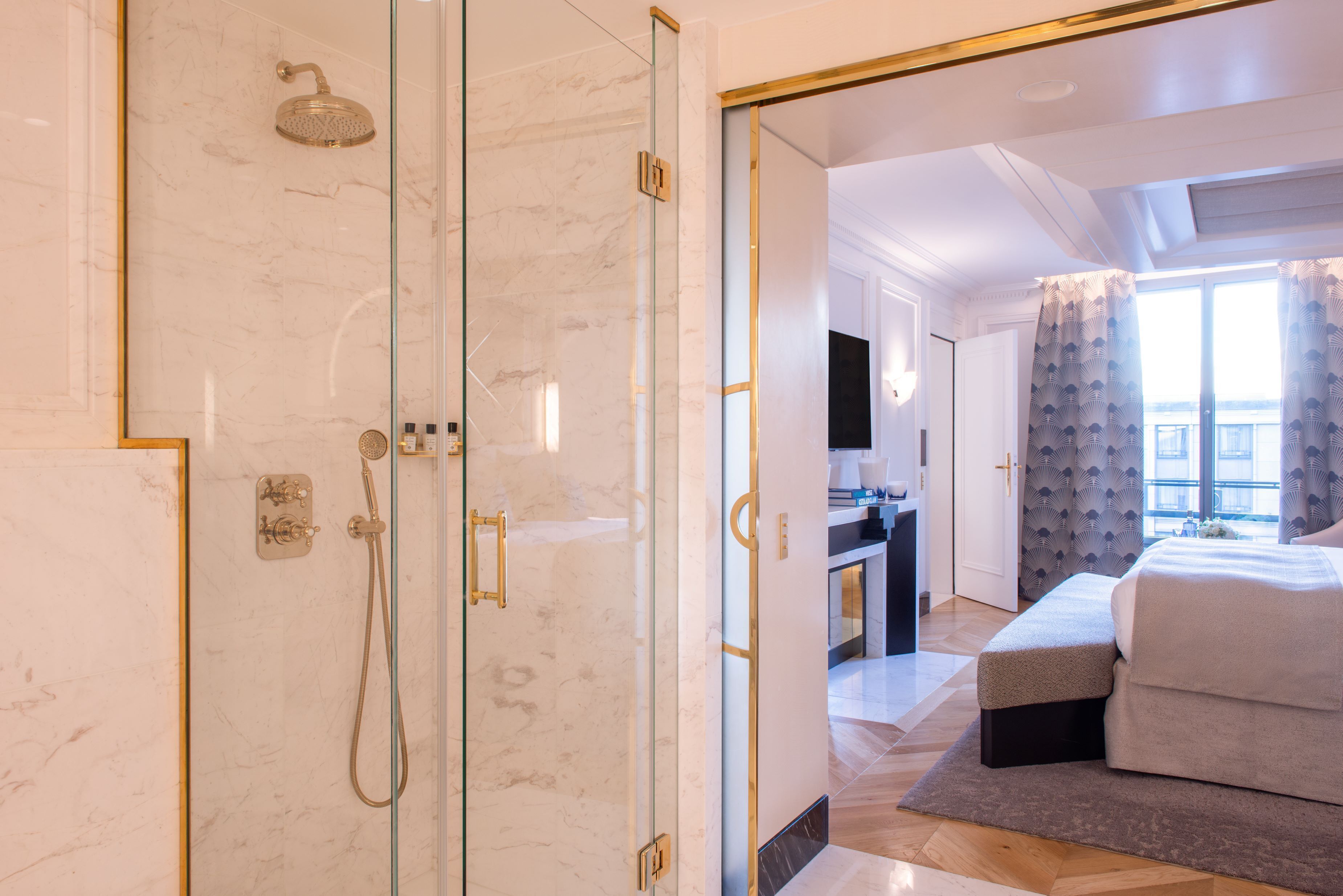 A modern hotel room with a glass-enclosed shower, a bed with a grey upholstered headboard, and a view of the city through large windows.