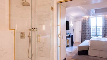 A modern hotel room with a glass-enclosed shower, a bed with a grey upholstered headboard, and a view of the city through large windows.