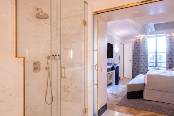 Una habitación de hotel moderna con ducha envuelta en vidrio, cama con cabecera tapizada en gris y vistas de la ciudad a través de amplias ventanas.