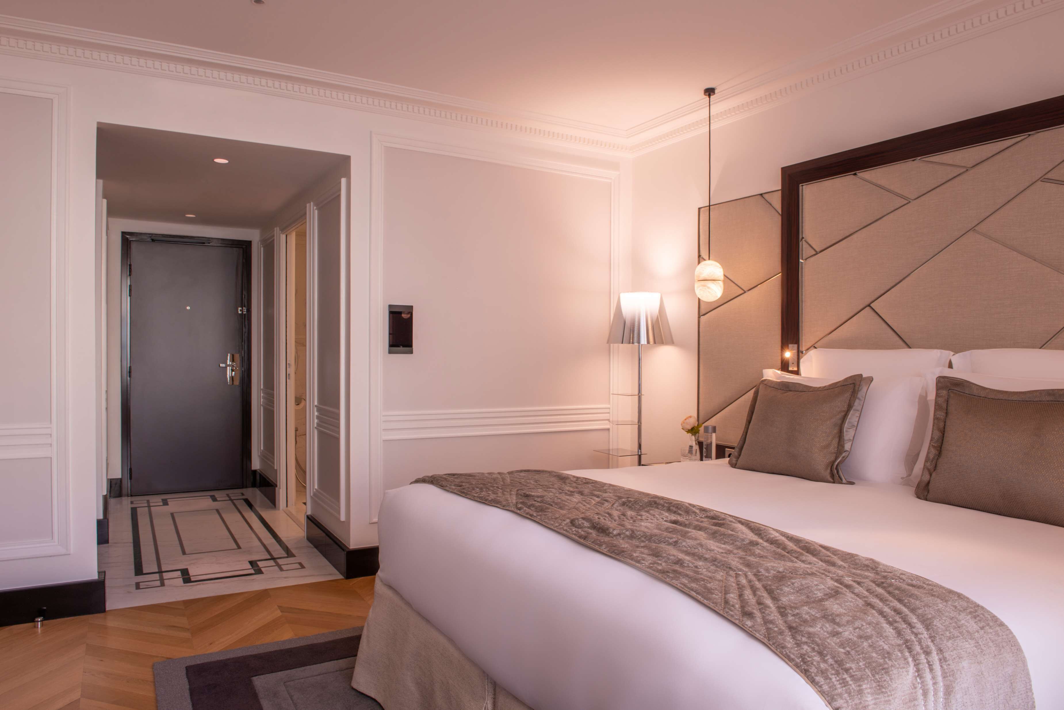 Habitación de hotel con una cama grande, una mesita de noche, una lámpara y una puerta que da a otra habitación.