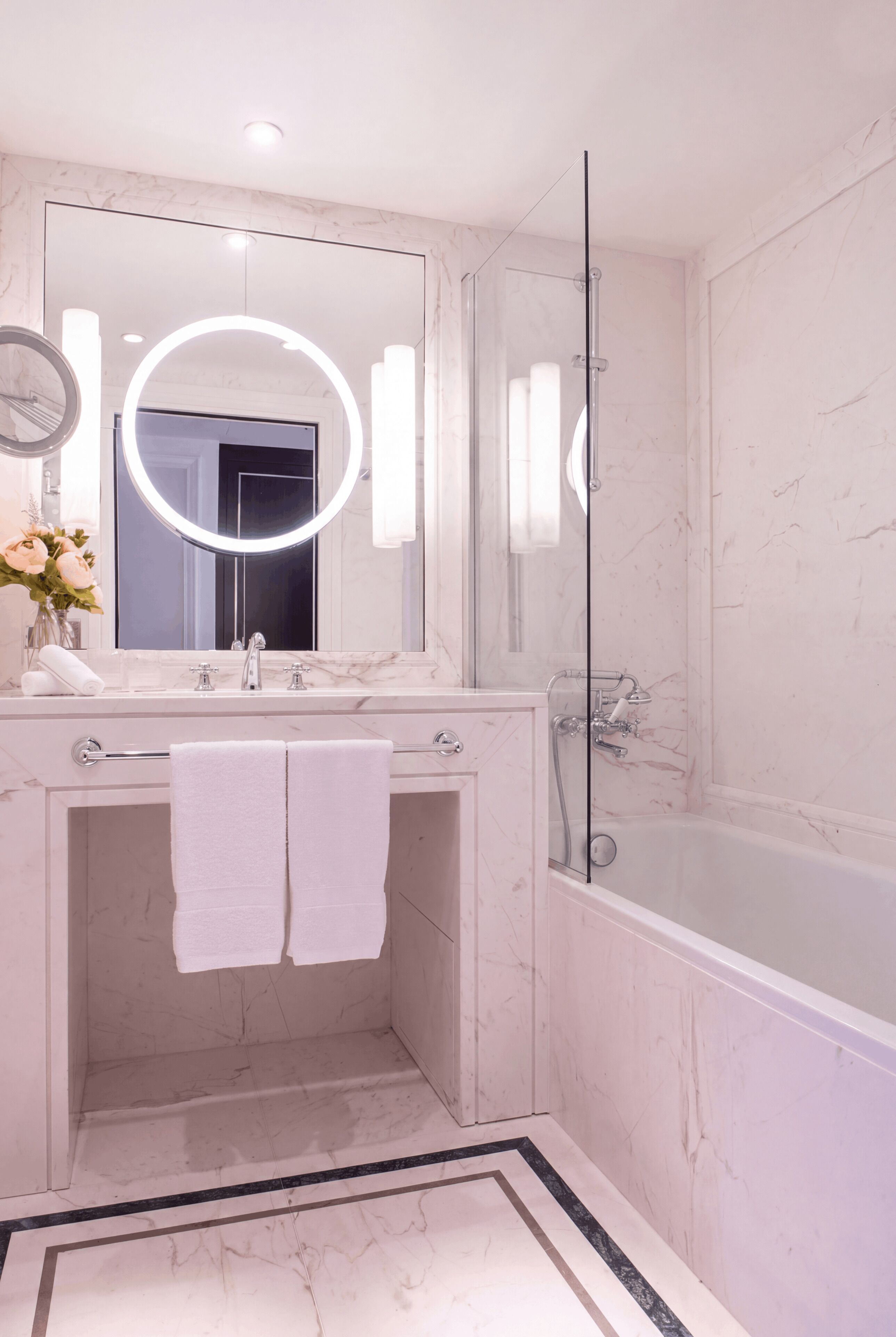 A bathroom with a marble vanity, a glass shower enclosure, and a large mirror with a circular light.