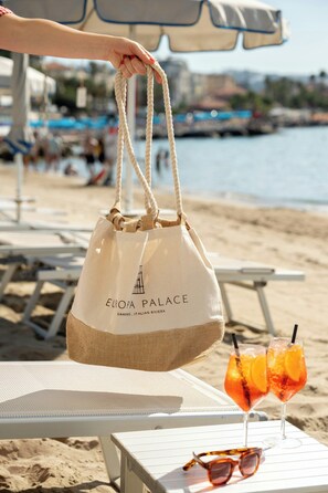 A beachside setting with a person holding a bag labeled 'EUROPA PALACE' and a table with drinks and sunglasses. - Hotel Europa (Sanremo)