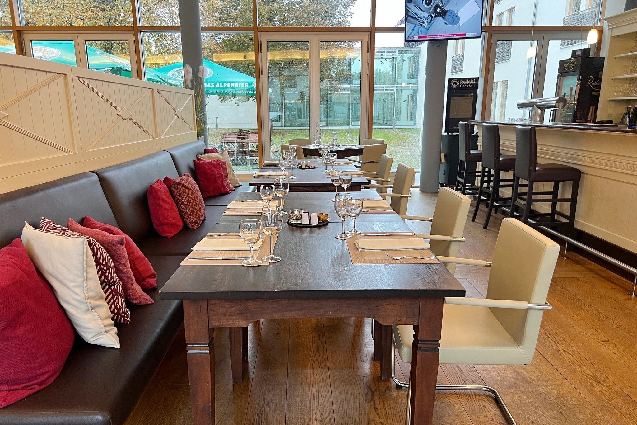 A restaurant with a long dining table set for a meal, a bar area with a TV, and a view of the outdoors through large windows.