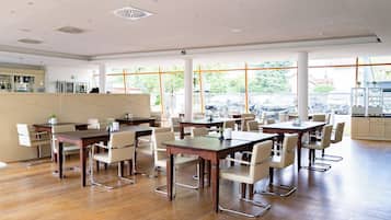 Un vaste espace dédié à la salle à manger, avec de nombreuses tables et chaises, de grandes fenêtres aux montures orange et une cuisine visible à travers ces fenêtres.