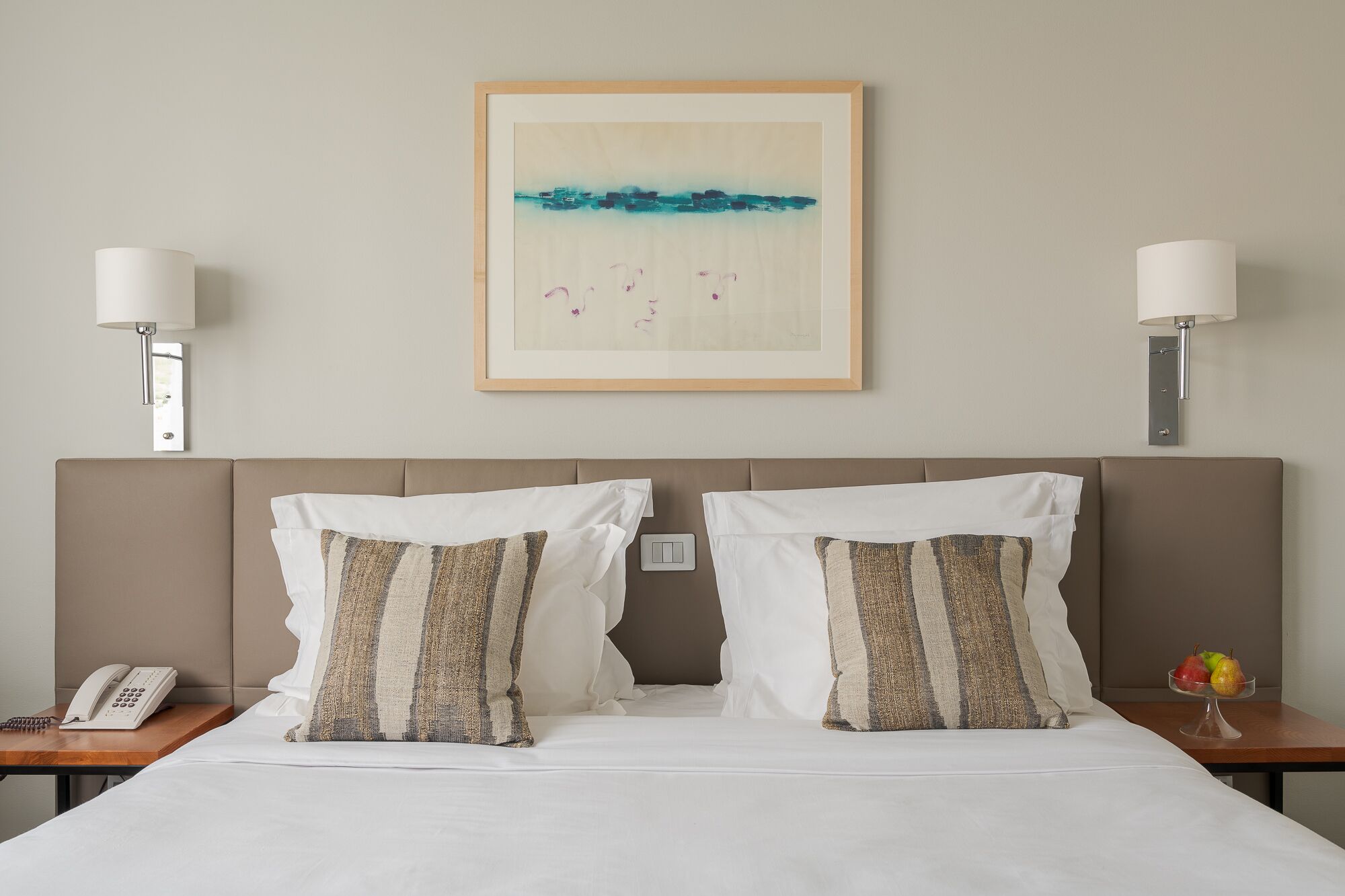 Een hotelkamer met een bed, twee bedlampen, een lijst met kunstwerk, een telefoon en een schaal fruit.