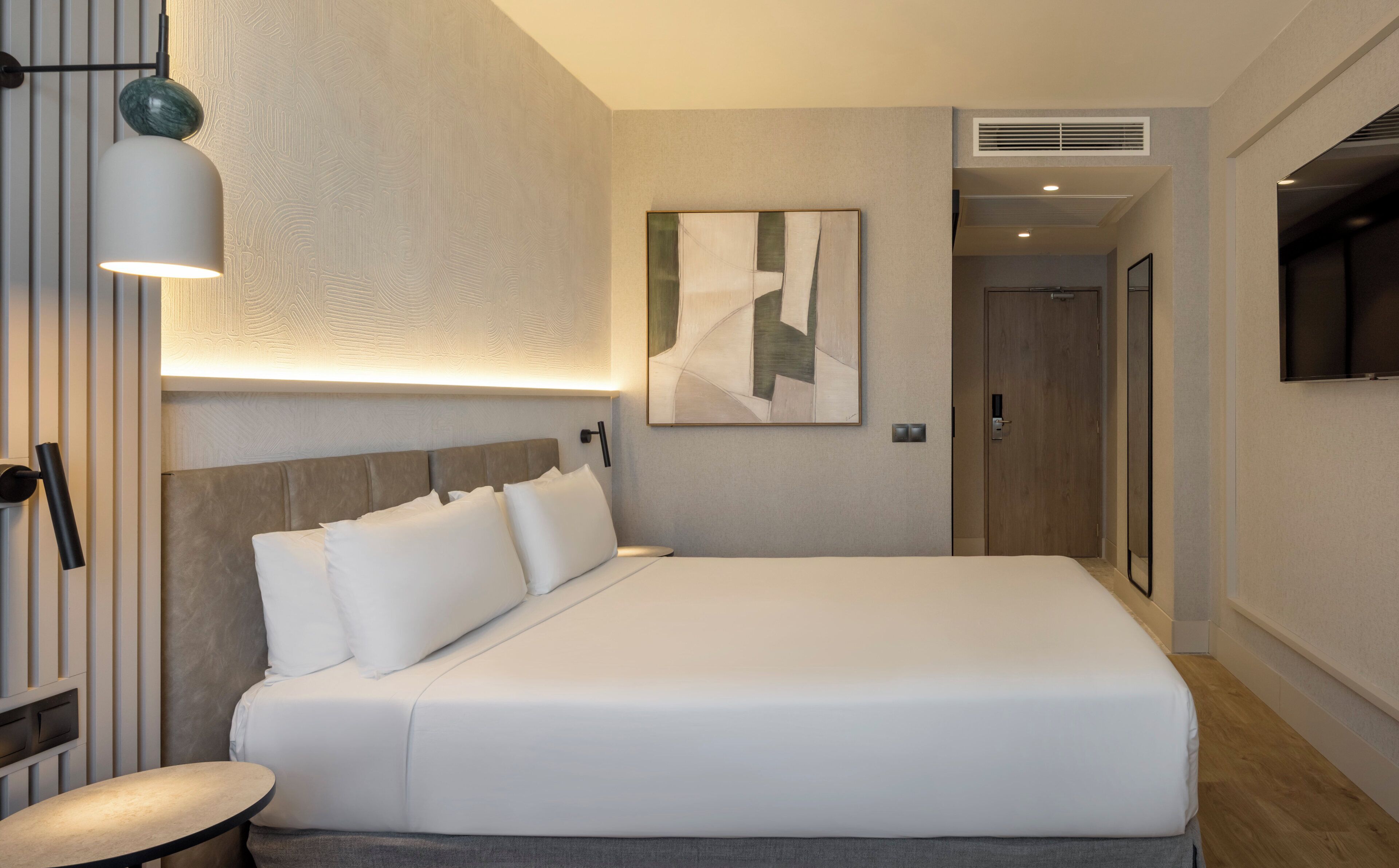 A modern hotel room with a large bed, a bedside table, a wall-mounted lamp, and a painting.