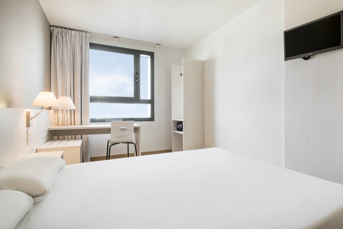 A hotel room with a bed, a desk, a chair, a TV, and a window with curtains.
