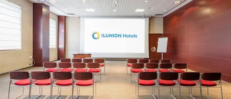 Un salón de conferencias con hileras de sillas rojas, una pantalla de proyector mostrando ILUNION Hotels y una pizarra.
