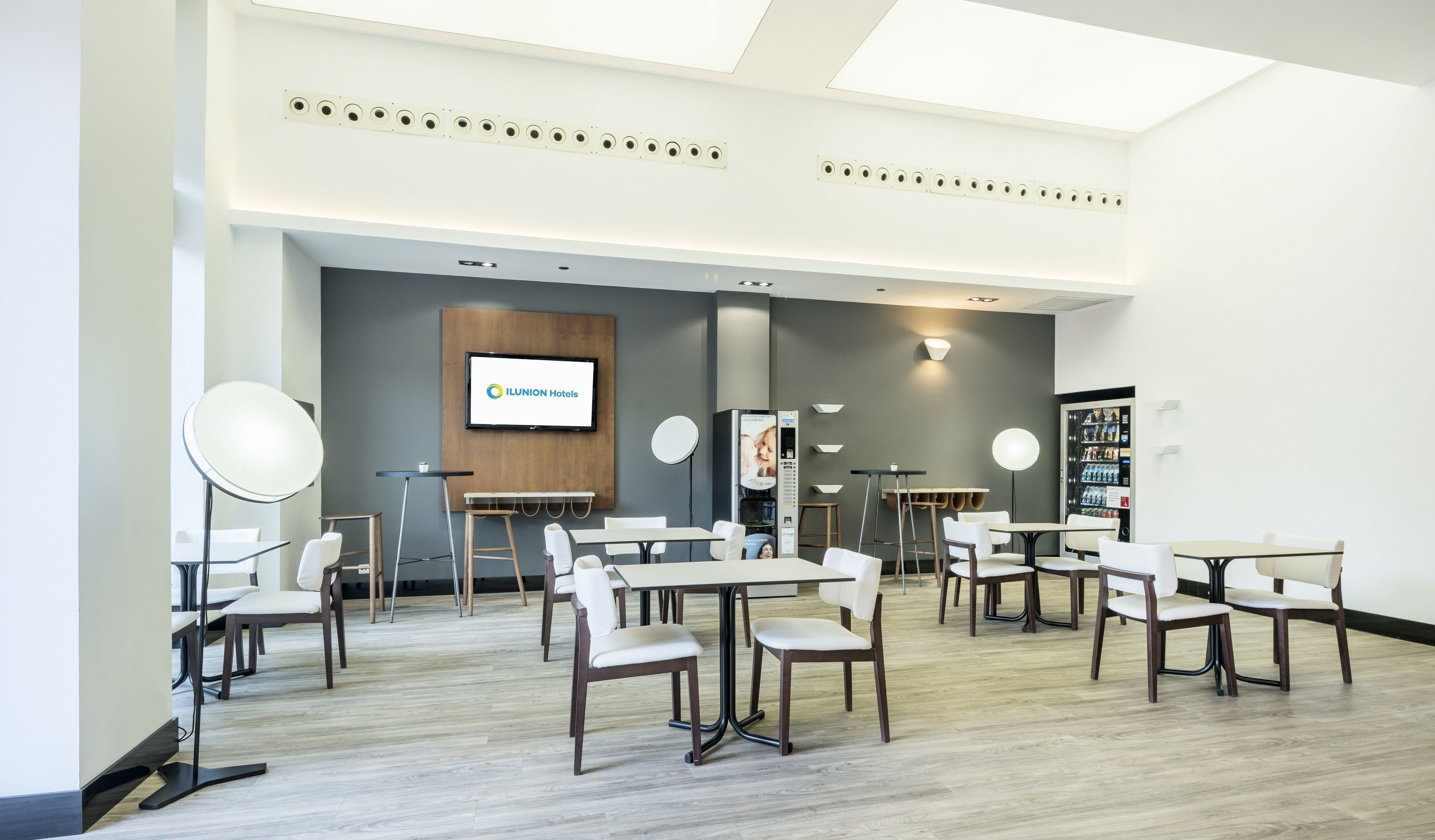A modern hotel conference room with multiple tables and chairs, a large screen displaying the hotel name, and a vending machine.