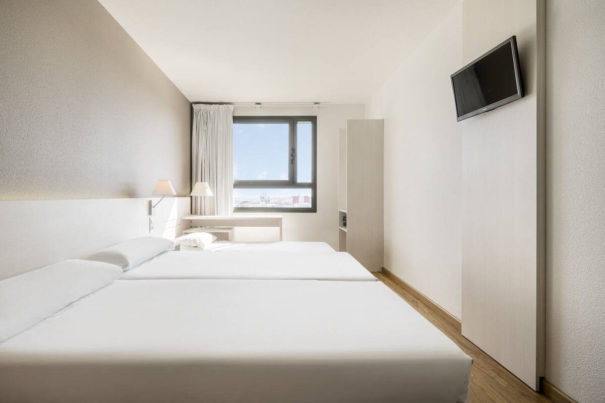 A hotel room with a bed, a desk, a lamp, a TV, and a window with a view of the city.