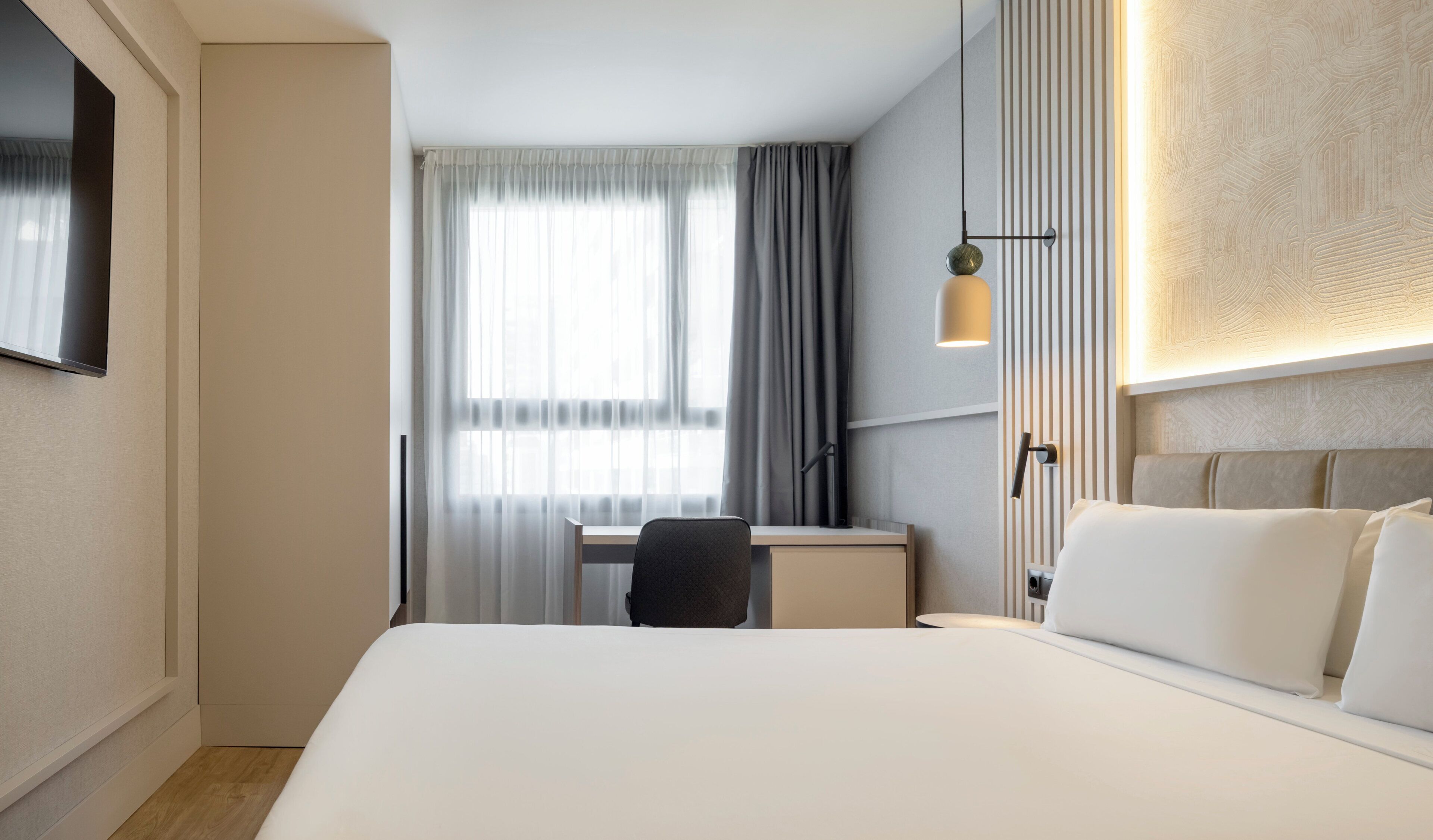 Ein Hotelzimmer mit Bett, Schreibtisch, Stuhl und einem an der Wand befestigten Fernseher.