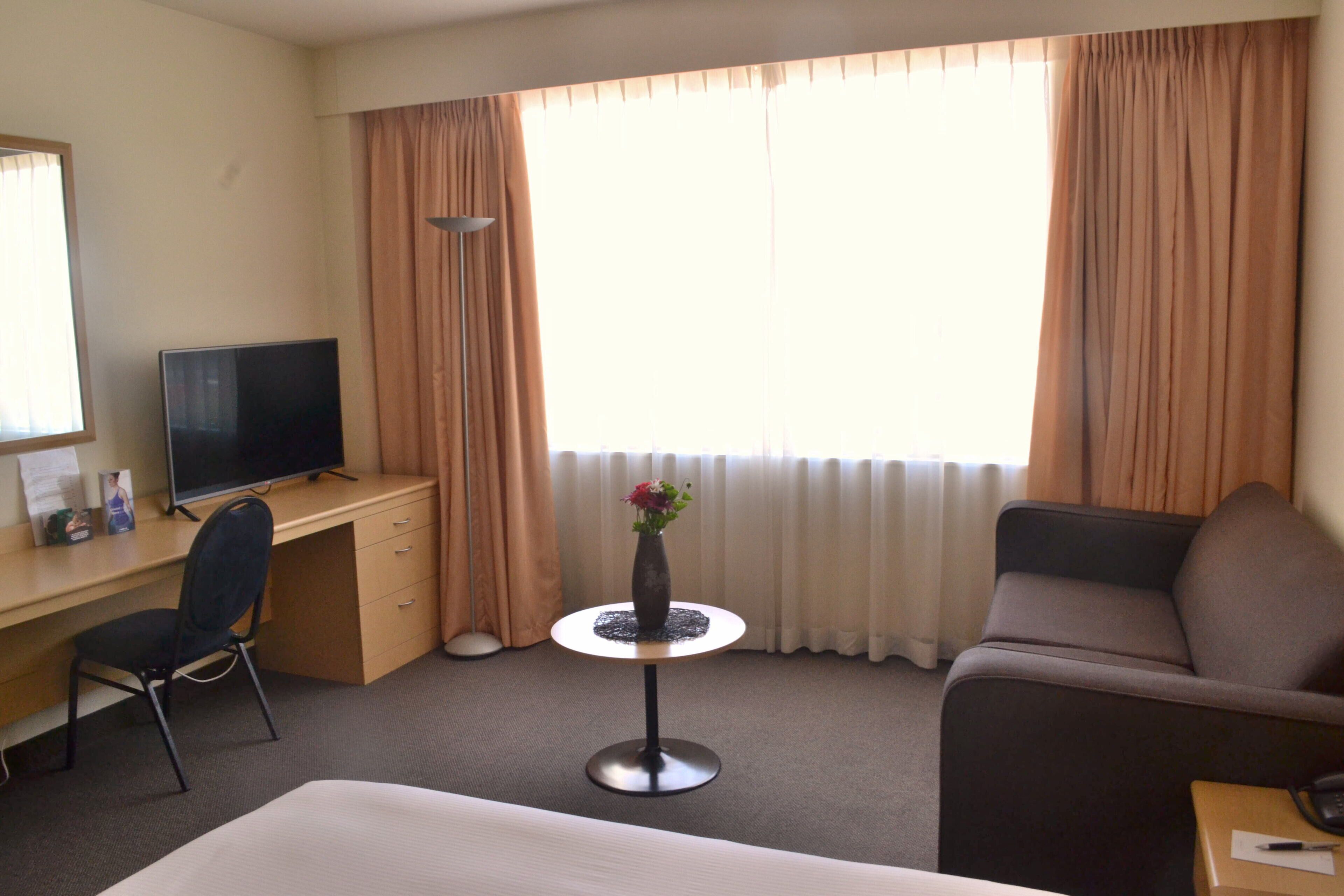 Een hotelkamer met een bed, een bureau met een televisie, een klein rond tafeltje met een vaas, een bank en gordijnen.