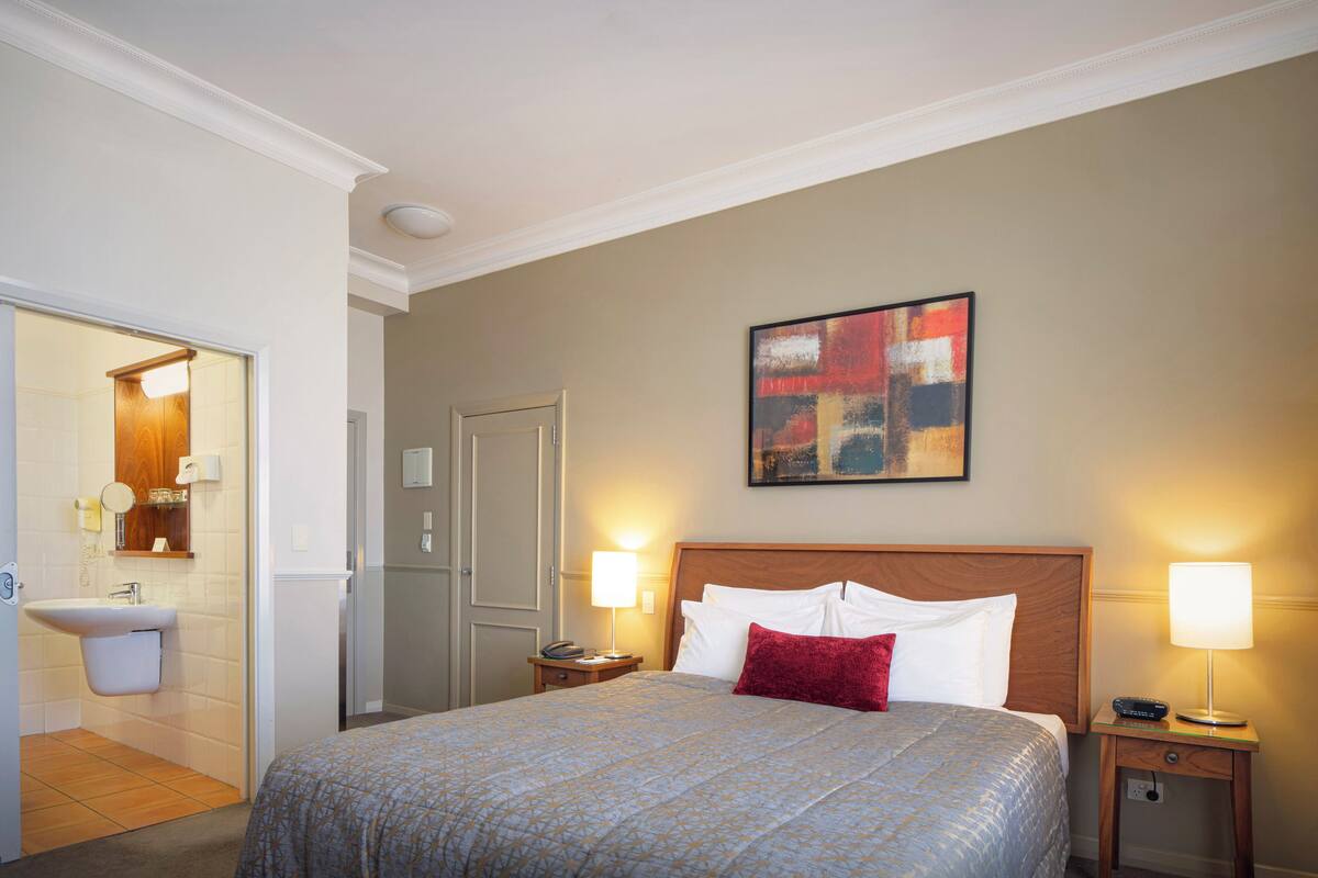 Una habitación de hotel con una cama, dos lámparas de noche, un cuadro en la pared y un baño visible a través de una puerta abierta.