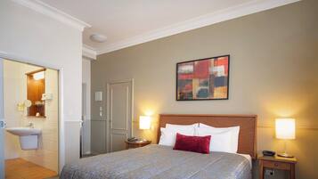 A hotel room with a bed, two bedside lamps, a painting on the wall, and a bathroom visible through an open door.