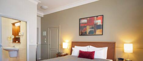 A hotel room with a bed, two bedside lamps, a painting on the wall, and a bathroom visible through an open door.