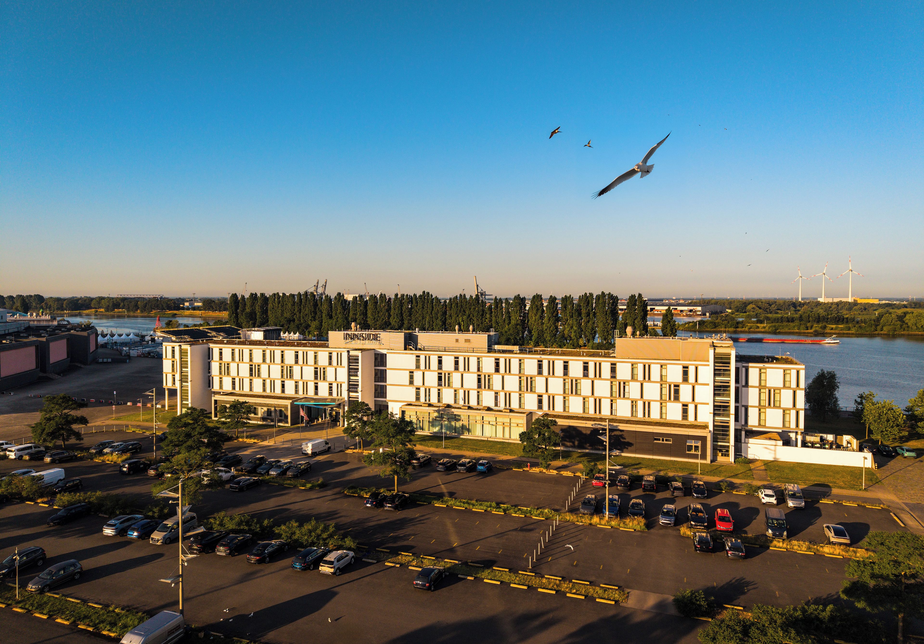 A multi-story hotel with a parking lot in front, situated near a body of water with wind turbines in the background.