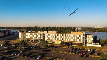A multi-story hotel with a parking lot in front, situated near a body of water with wind turbines in the background.