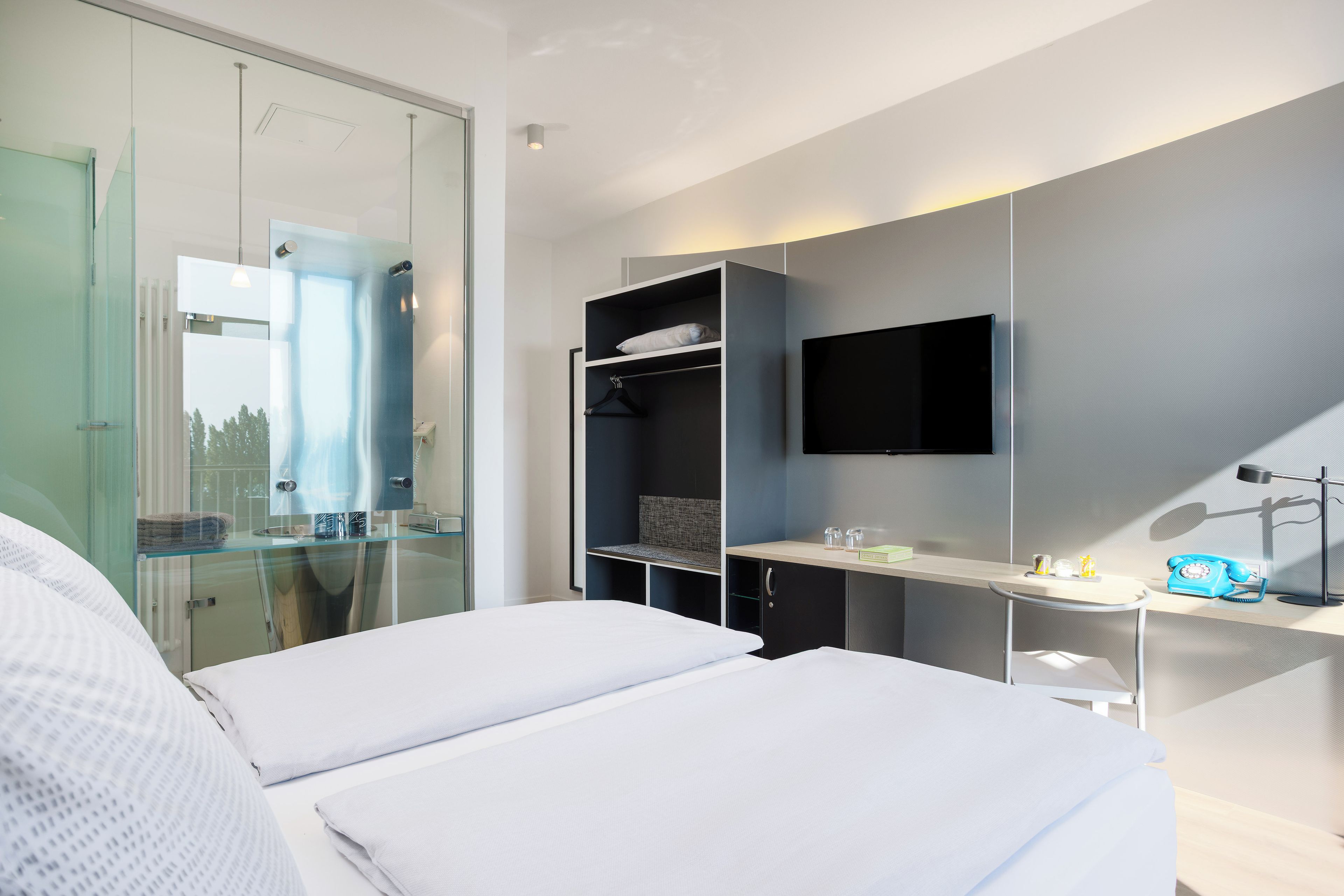 A modern hotel room with a bed, a desk with a computer, a TV, and a glass shower enclosure.