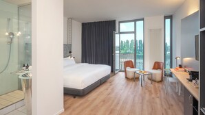 A modern hotel room with a large bed, a glass-enclosed shower, a balcony with a view, and a small dining area. - INNSiDE by Meliá Bremen (Bremen)