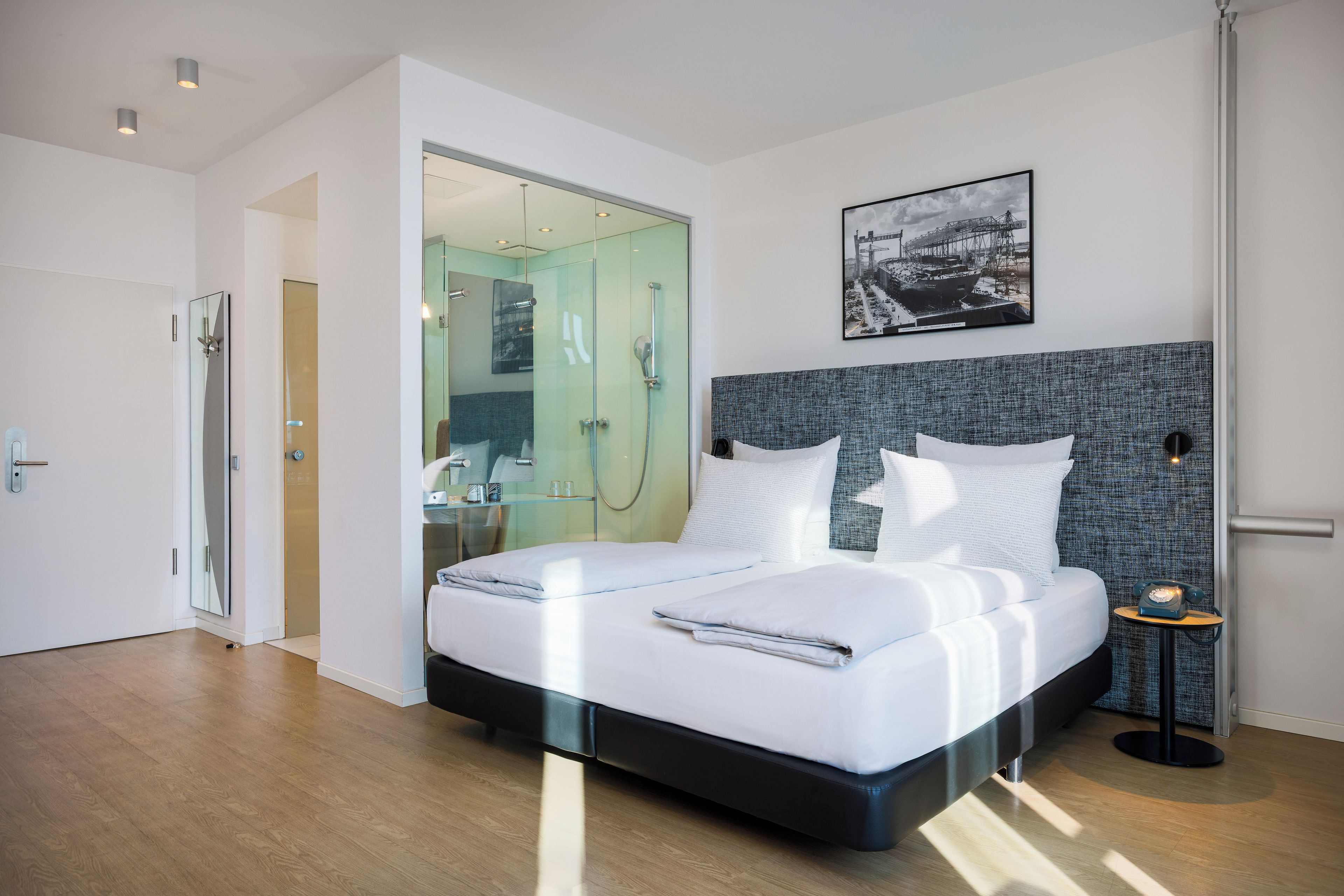 A modern hotel room with a large bed, a glass shower enclosure, and a view of the city.