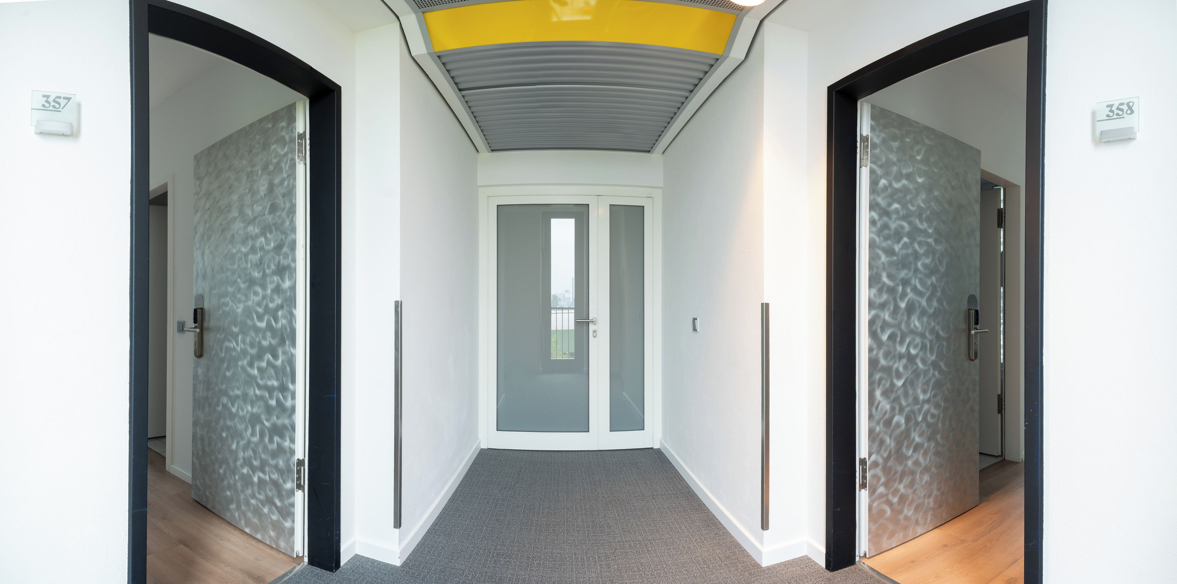 Un pasillo con diseño moderno, que presenta una pared texturizada, una puerta de cristal al final y un techo con detalles amarillos.
