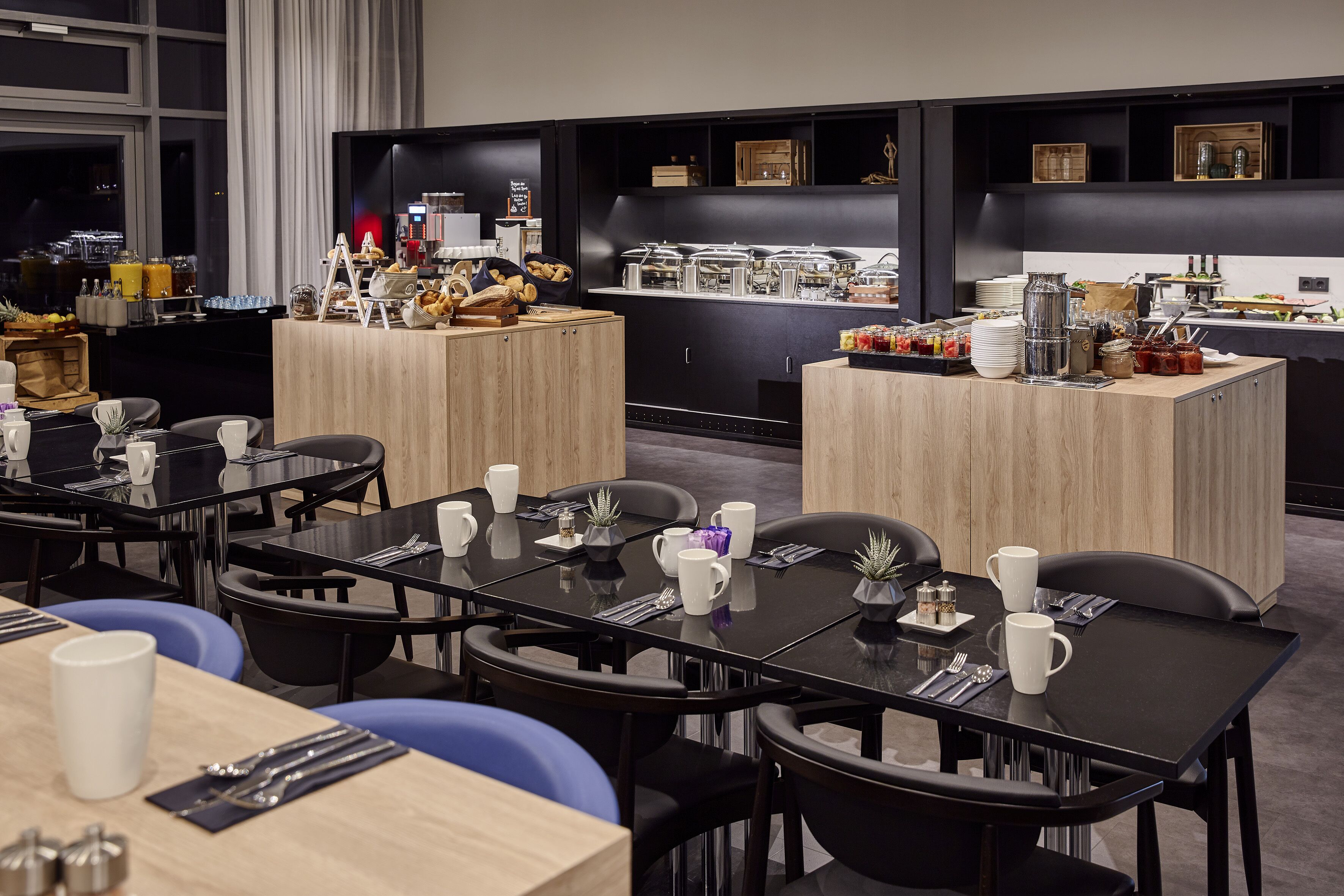 A modern dining area with black tables and chairs, a buffet counter with various food items, and a city view through large windows.