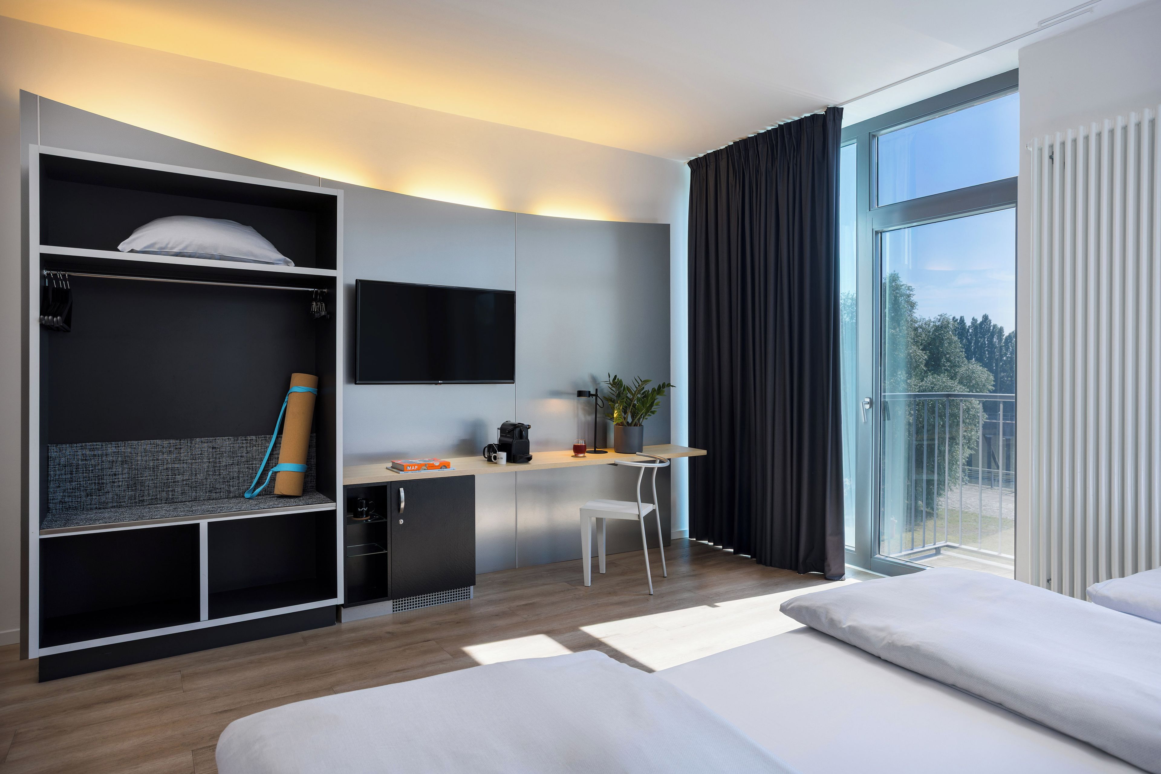 Ein modernes Hotelzimmer mit Bett, Schreibtisch, Stuhl, Kleiderschrank, Fernseher und einem Balkon mit Vorhängen.
