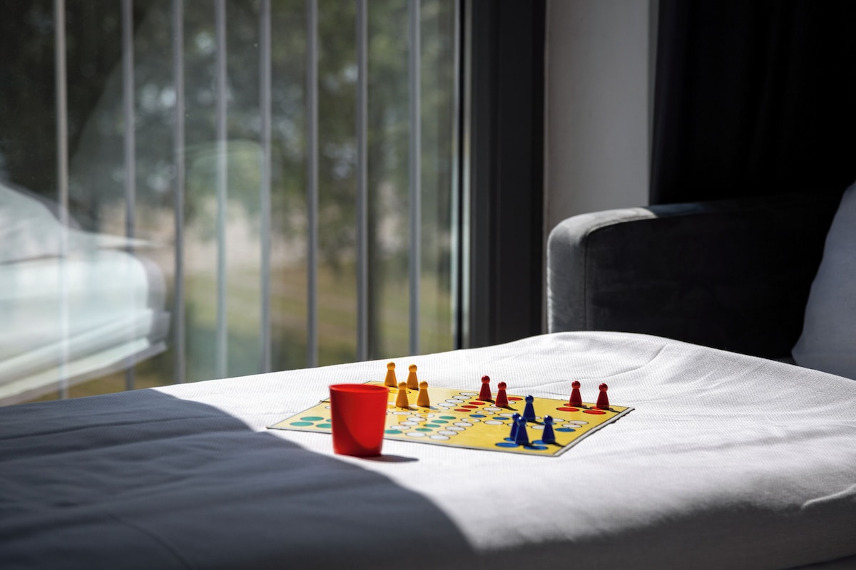 A bed with a chessboard and a red cup on it, near a window with blinds.
