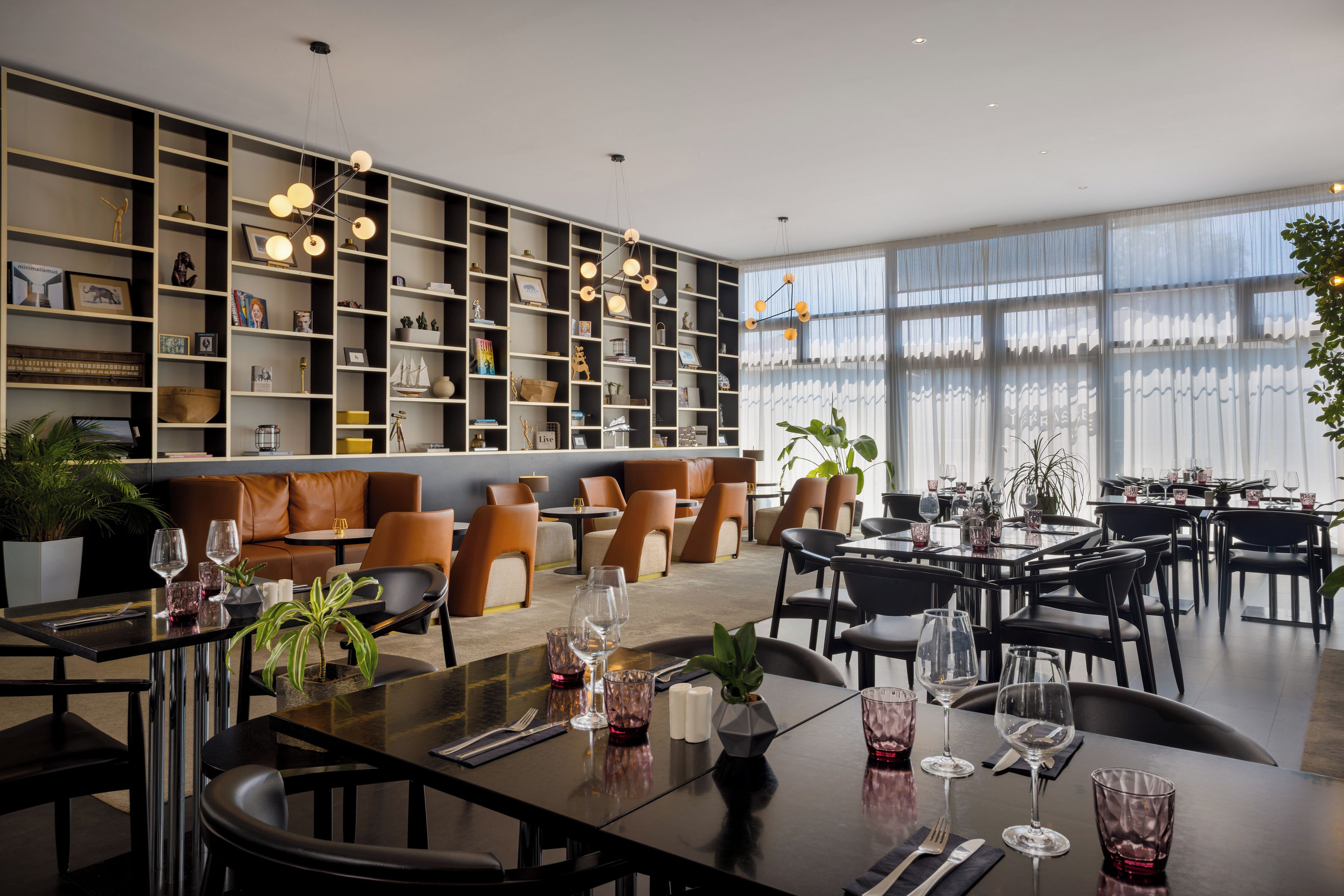 A modern dining area with a long communal table and individual seating, surrounded by a large bookshelf and a seating area with sofas.