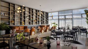 A modern dining area with a long communal table and individual seating, surrounded by a large bookshelf and a seating area with sofas.