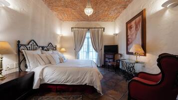 A bedroom with a brick ceiling, a bed with white linens, a red chair, a desk with a lamp, and a painting on the wall.