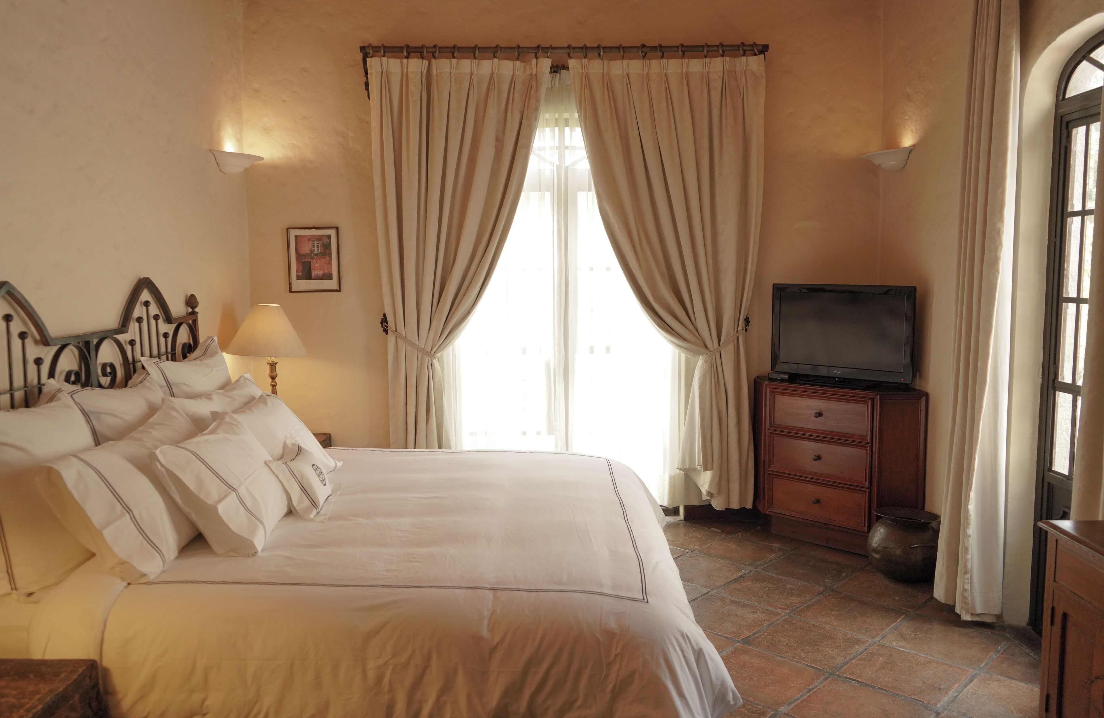 A bedroom with a bed, a TV on a wooden cabinet, and a window with curtains.