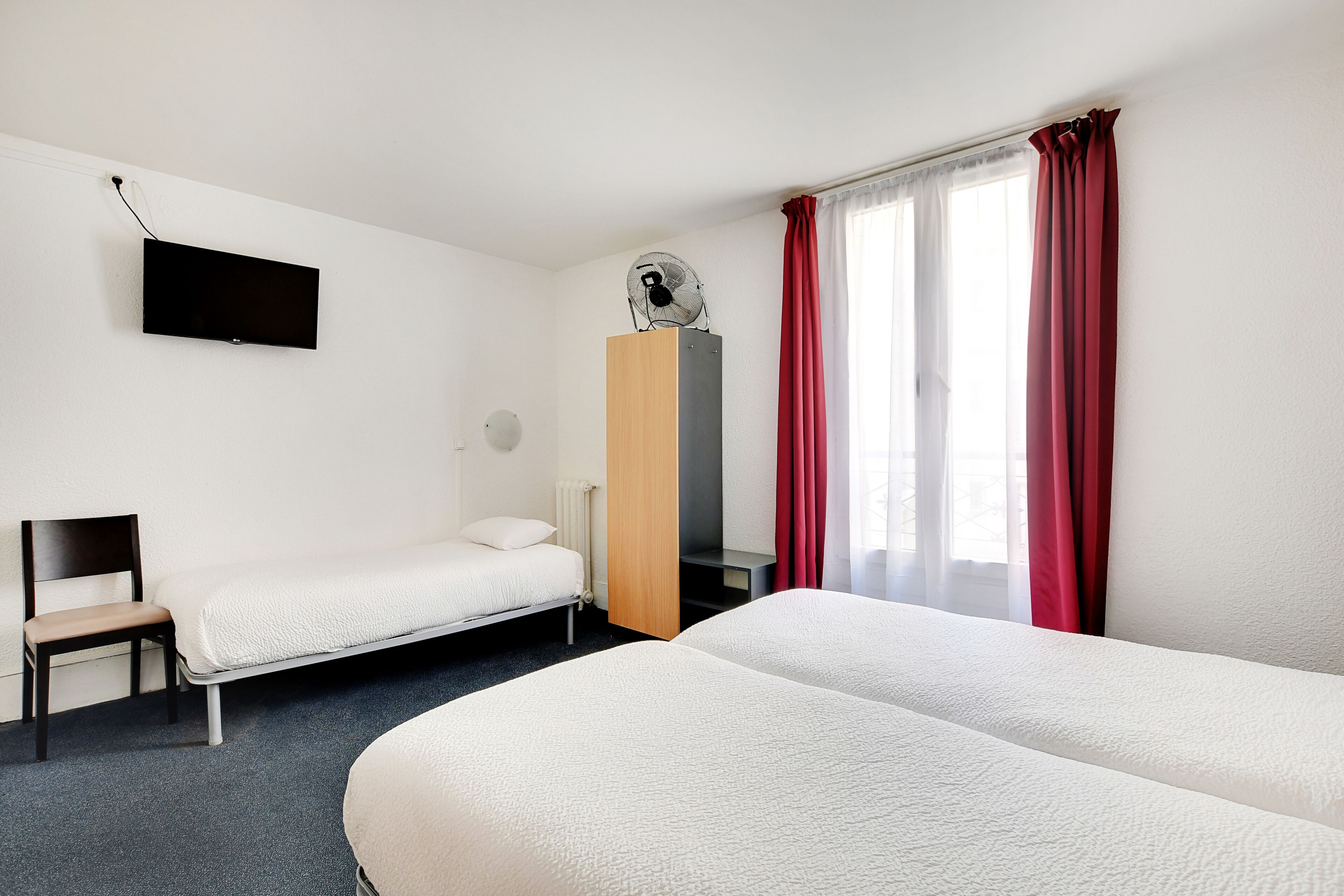 A hotel room with two beds, a chair, a TV mounted on the wall, a standing fan, and a window with red curtains.