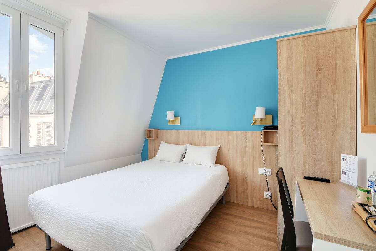 A bedroom with a bed, a desk, and a chair. There is a window with blinds, a wall-mounted lamp, and a wooden headboard.