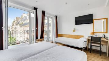 Ein Hotelzimmer mit zwei Betten, einem Schreibtisch, einem Stuhl, einem Fernseher und einem Balkon mit Blick auf Gebäude.