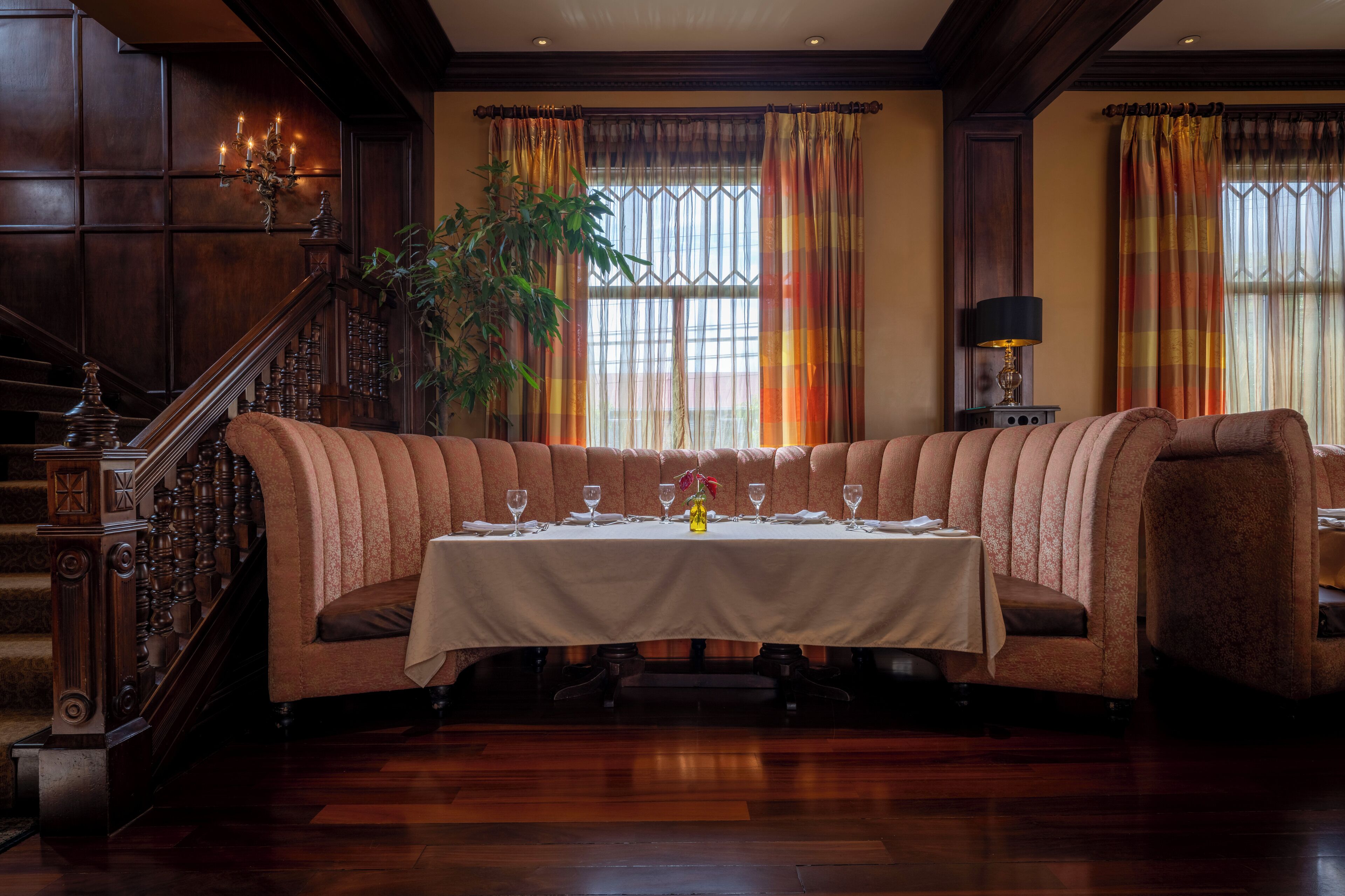Un comedor formal con una larga mesa dispuesta para seis, una escalera con barandales ornamentales y grandes ventanales con cortinas de colores.