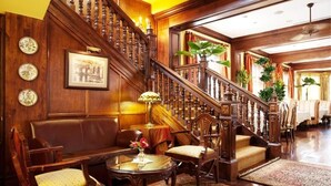 A grand staircase with wooden railings and a chandelier, leading to a room with a sofa, chairs, and a table. - Hotel Grano de Oro (San José)