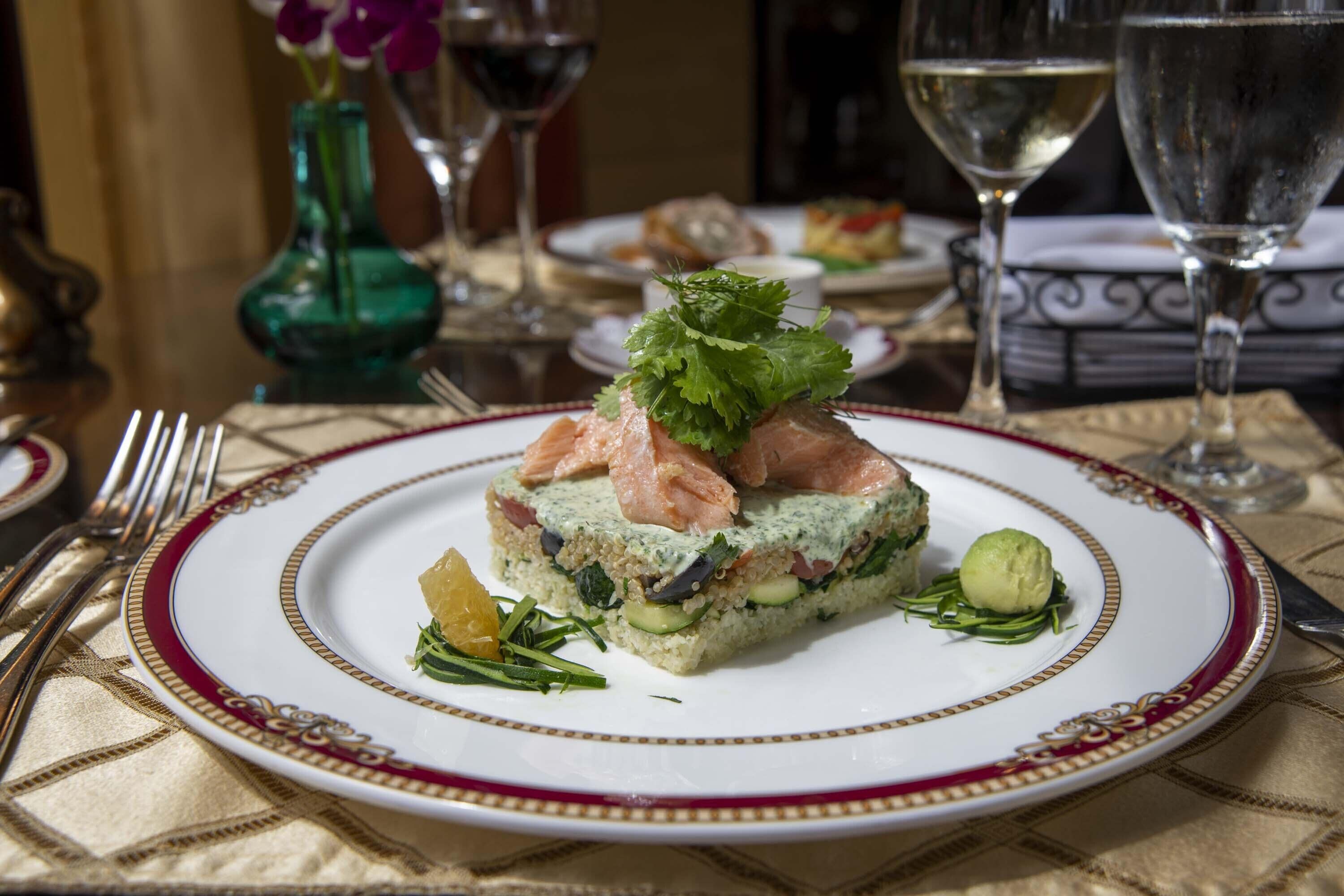 Une assiette de nourriture ornée d’un bord décoratif, un verre de vin blanc et un vase vert en arrière-plan.