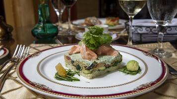Une assiette de nourriture ornée d’un bord décoratif, un verre de vin blanc et un vase vert en arrière-plan.
