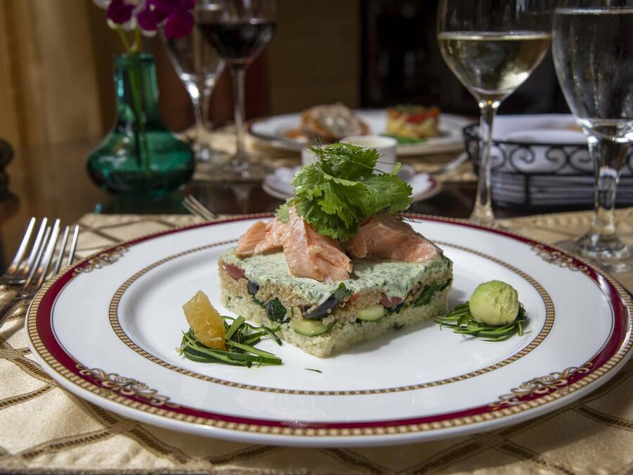 Une assiette de nourriture ornée d’un bord décoratif, un verre de vin blanc et un vase vert en arrière-plan.
