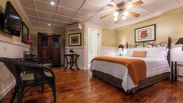 A room with a bed, a ceiling fan, a TV, a wooden floor, and a dining table with chairs.
