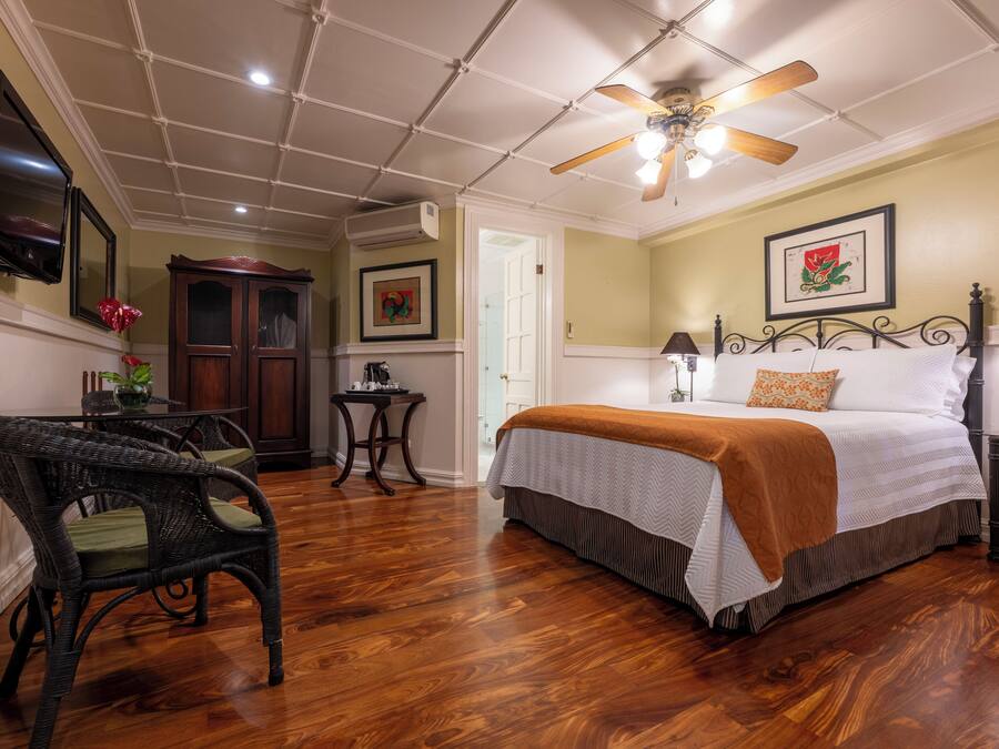 A room with a bed, a ceiling fan, a TV, a wooden floor, and a dining table with chairs.