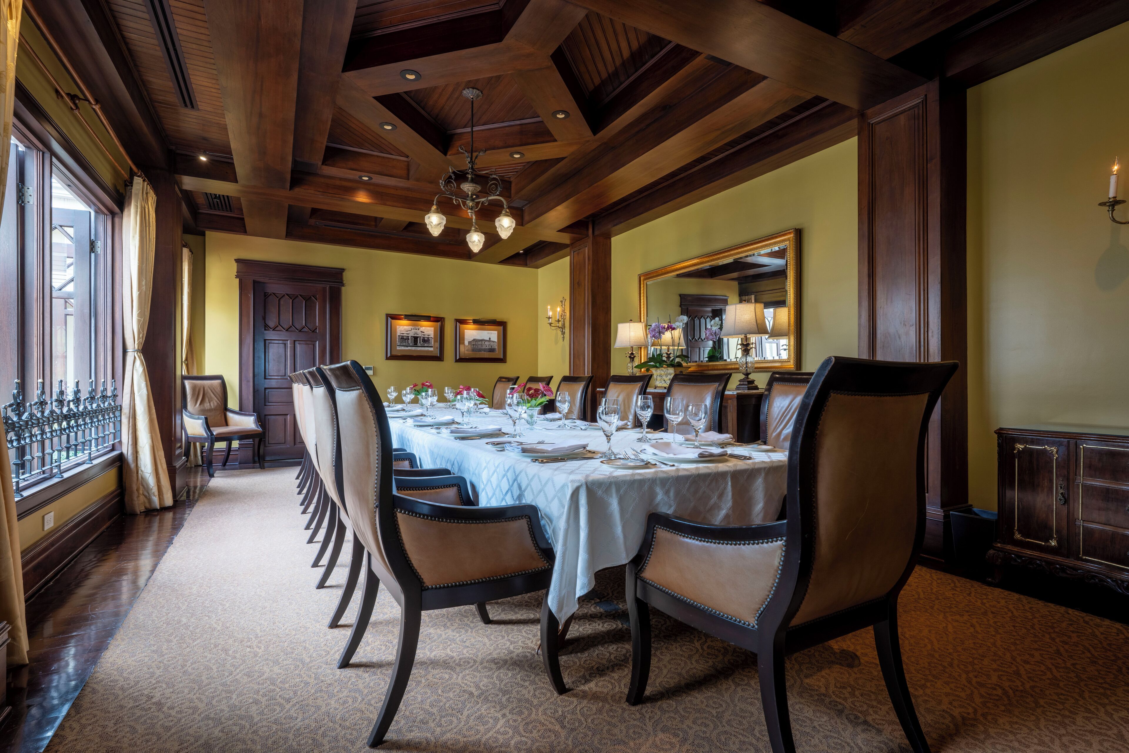 Uma sala de jantar formal com uma mesa comprida posta para refeição, vigas de madeira no teto e um lustre.