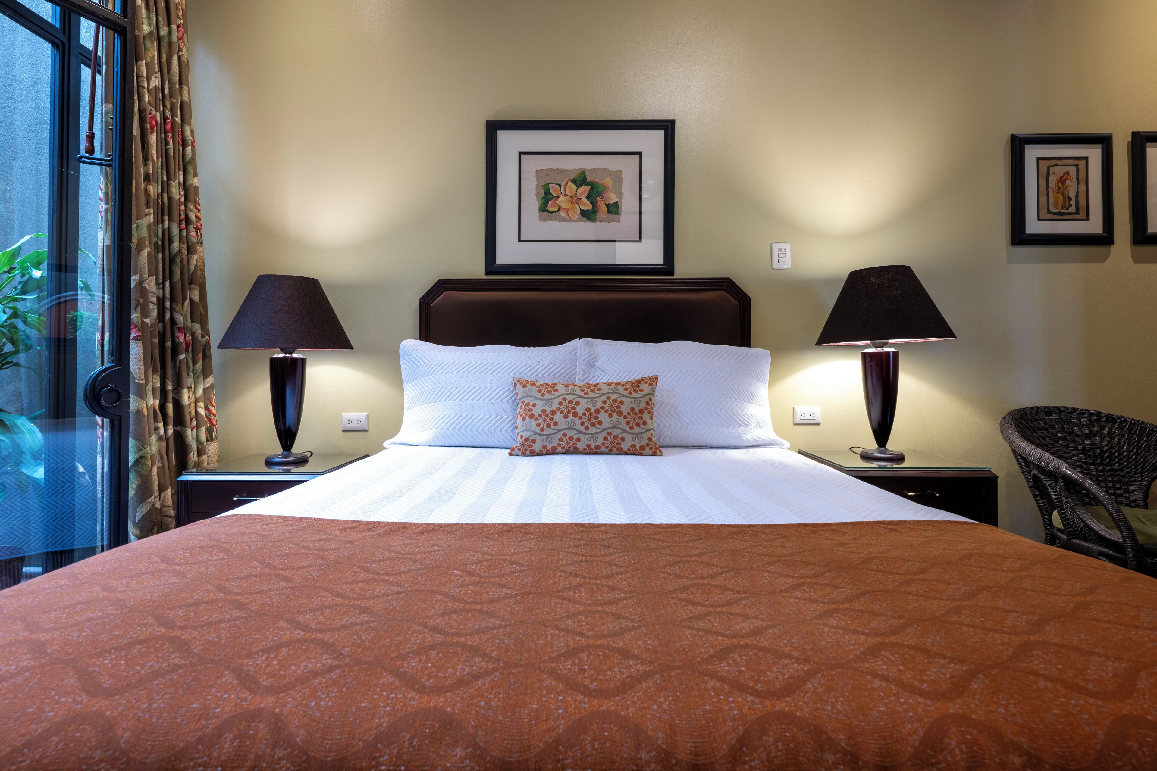 A hotel room with a large bed, two bedside lamps, a framed picture on the wall, and a wicker chair.