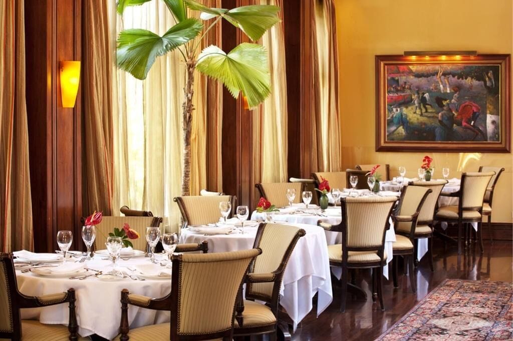 A dining area with tables set for a meal, chairs, and a large painting on the wall.