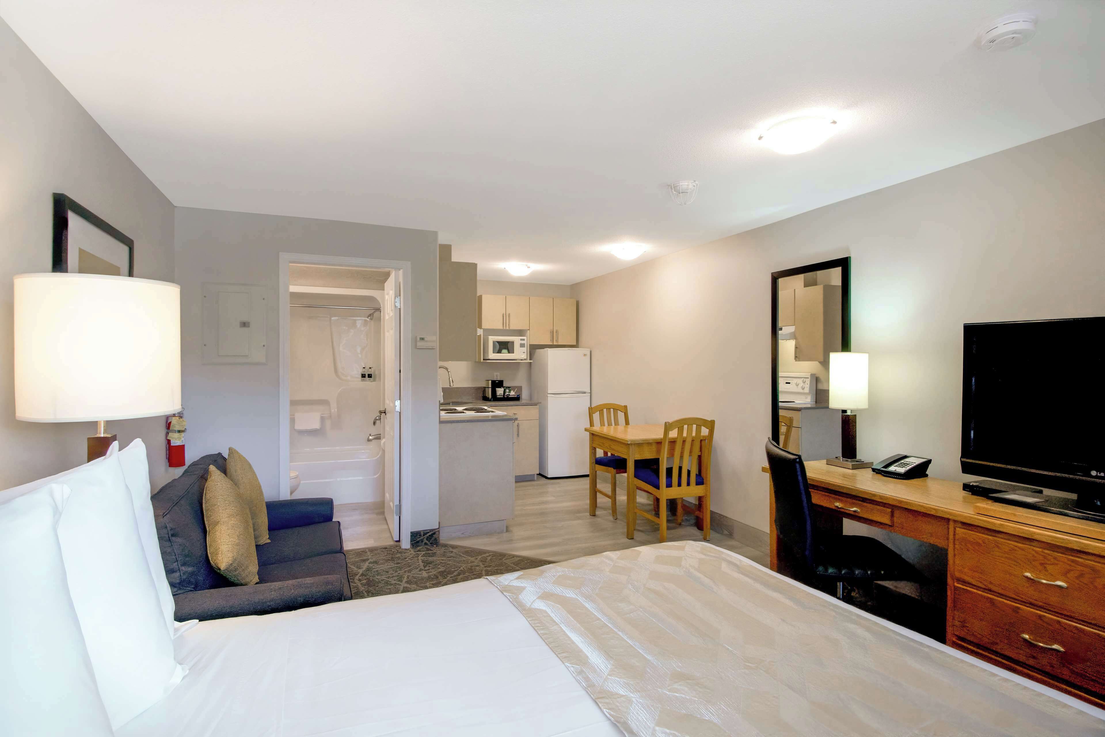 Habitación de hotel con cama, escritorio, silla, televisión y una kitchenette al fondo.