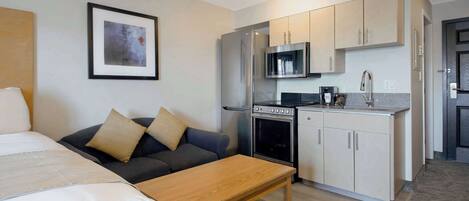 A compact hotel room with a bed, a sofa, a wooden table, a kitchenette with a fridge, microwave, stove, and cabinets.
